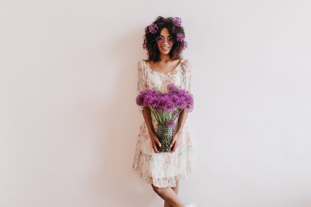 Dama africana glamorosa en vestido de verano con jarrón de flores. Toma interior de mujer rizada inspirada disfrutando de una sesión de fotos en casa.