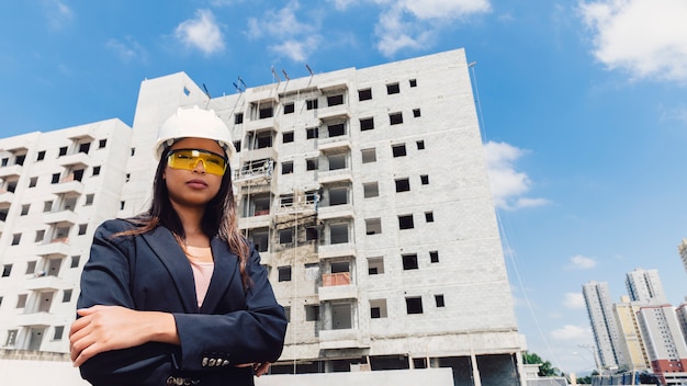 Dama afroamericana en casco de seguridad y anteojos cerca del edificio en construcción