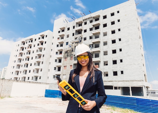 Dama afroamericana en casco de seguridad con teléfono inteligente y nivel de edificio cerca del edificio en construcción