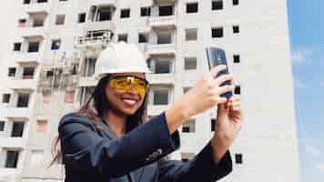 Foto gratuita dama afroamericana en casco de seguridad tomando selfie cerca de edificio en construcción