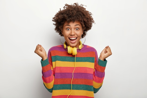 Una dama afroamericana extremadamente feliz celebra el gol anotado, hace un gesto de victoria, levanta los puños cerrados, tiene un peinado rizado, usa un jersey de rayas de colores, usa auriculares conectados a algún dispositivo