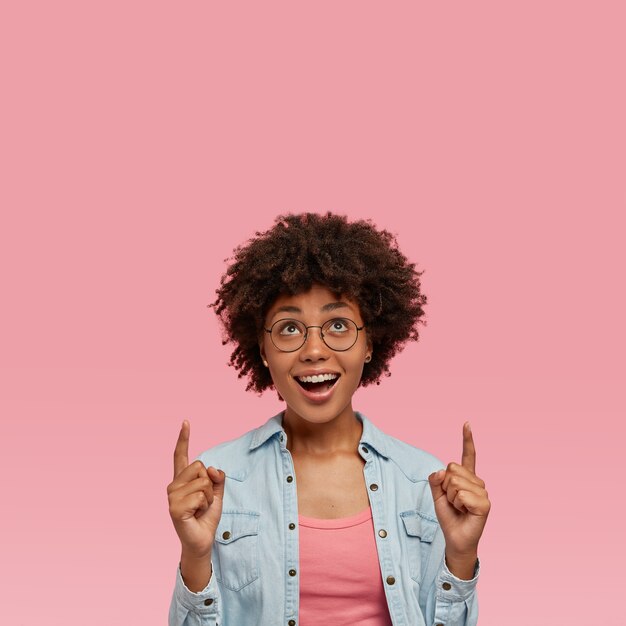 La dama afroamericana de moda tiene el cabello oscuro y rizado, usa anteojos redondos, indica con ambos dedos índices hacia arriba, muestra la dirección en el piso de arriba, feliz de comprar un apartamento nuevo, aislado en la pared rosa