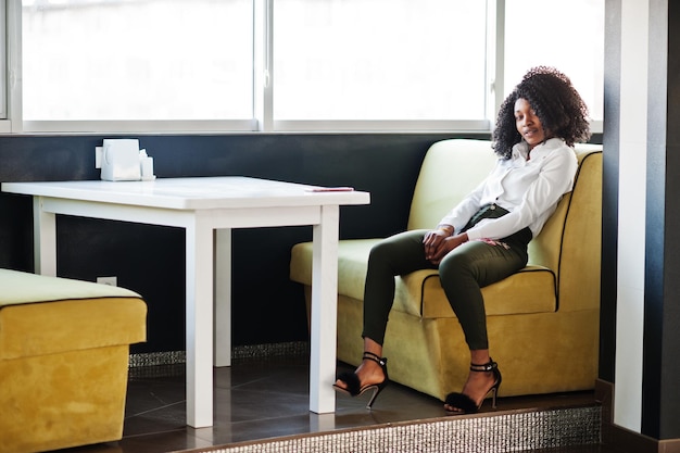 Una dama afroamericana de negocios pura con cabello afro usa blusa blanca y pantalones verdes posados en un café