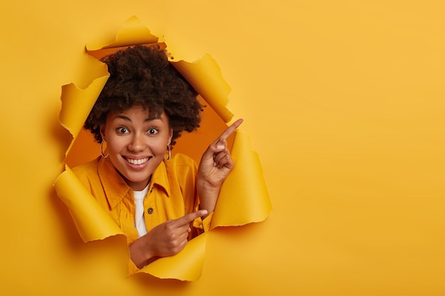 La dama afroamericana positiva muestra algo sorprendente, apunta hacia arriba y hacia los lados con los dedos índices, anuncia espacio de copia, tiene una sonrisa con dientes, aislado sobre fondo amarillo