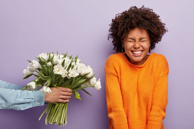 La dama afroamericana rizada joven encantadora llena de alegría recibe felicitaciones y flores en el cumpleaños, el hombre irreconocible extiende las manos y da tulipanes blancos de primavera, aislados sobre la pared púrpura.