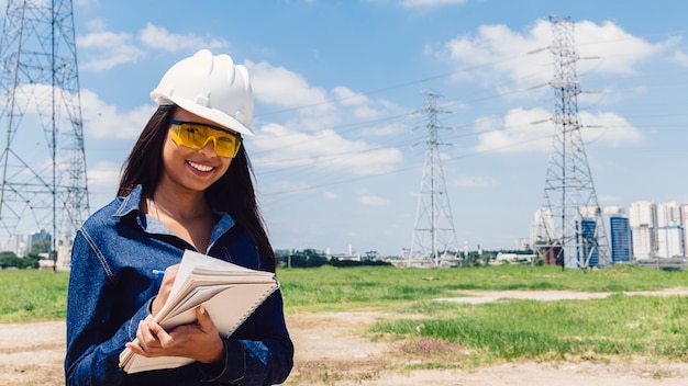 Dama afroamericana sonriente en casco de seguridad tomando notas cerca de línea de alto voltaje