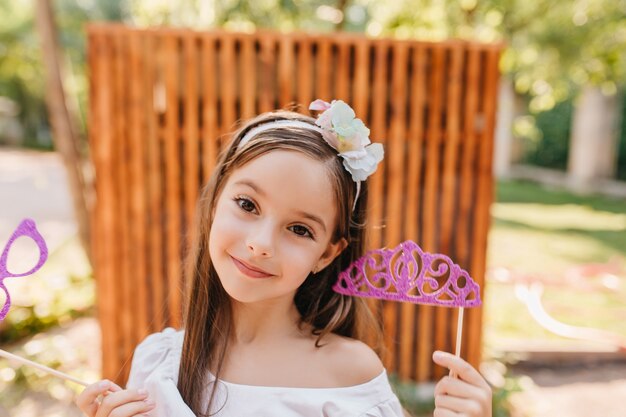 Dama alegre con grandes ojos marrones sosteniendo una corona de juguete rosa y posando en el patio con placer. Retrato de primer plano al aire libre de una niña morena sonriente con gafas de brillo en la mano.