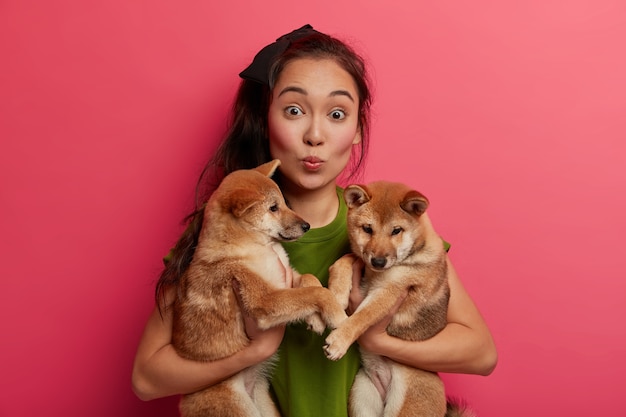 Foto gratuita dama asiática sorprendida mantiene los labios doblados, posa con dos cachorros y los lleva a la clínica veterinaria.