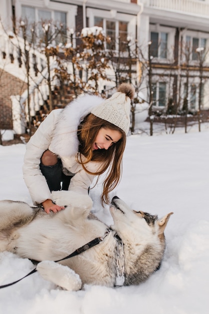 Dama blanca inspirada con sombrero jugando con husky en la nieve. Foto exterior de risa joven jugando con su perro en el patio.