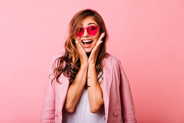 Dama blanca refinada posando con sonrisa sorprendida en rosa. Hermosa chica de pelo largo con divertidas gafas rosas jugando.