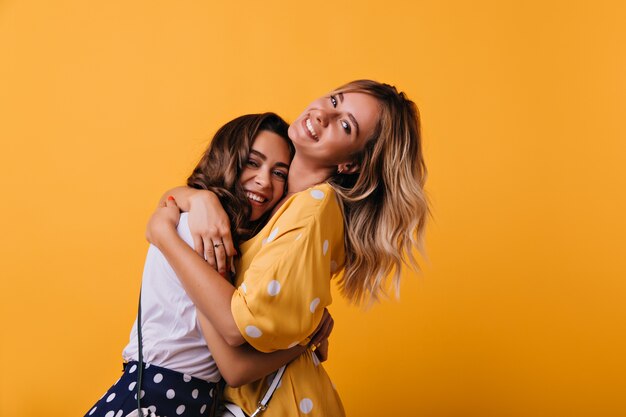 Dama blanca romántica en vestido amarillo abrazando a su hermana. Retrato interior de una hermosa chica de moda relajándose con su mejor amigo en la sesión de retratos de fin de semana.