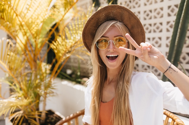 Dama bronceada de ensueño con gafas de sol amarillas jugando en el café del complejo. Retrato al aire libre de la adorable chica rubia posando con una sonrisa de sorpresa.