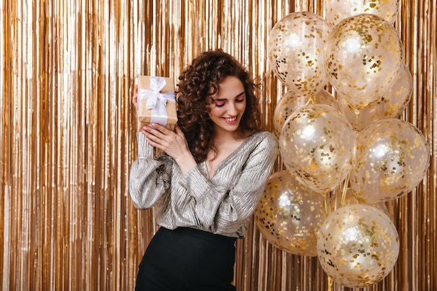 Foto gratuita dama con cabello ondulado sacudiendo caja de regalo sobre fondo dorado