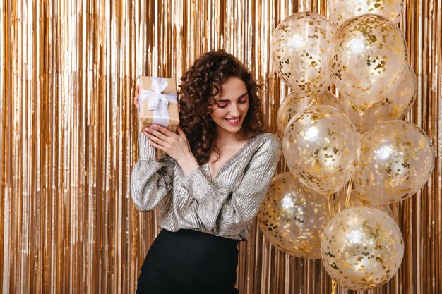 Dama con cabello ondulado sacudiendo caja de regalo sobre fondo dorado