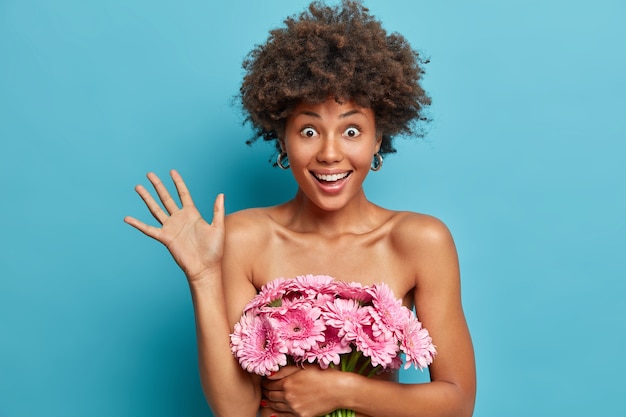 La dama de cabello rizado emocional llena de alegría sostiene un ramo de flores, levanta la palma, cubre el cuerpo desnudo, tiene ojos saltones y una sonrisa con dientes, piel oscura bien cuidada