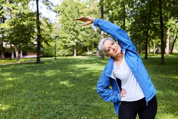 Dama caucásica senior de cabello gris activa sosteniendo una mano en la cintura y levantando el brazo mientras hace curvas laterales en el parque, calentando el cuerpo antes del entrenamiento cardiovascular, teniendo una expresión facial feliz y alegre