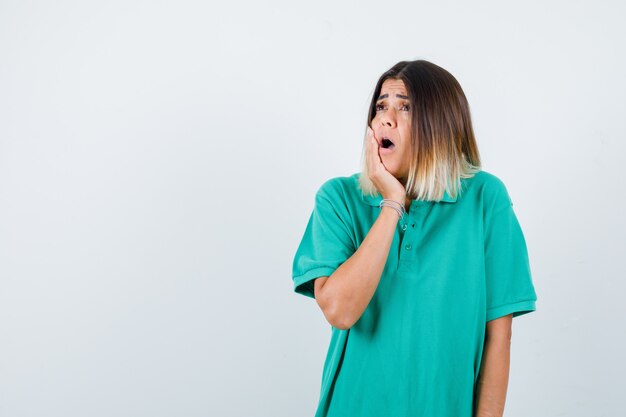 Dama elegante en camiseta de polo con la mano en la mejilla, mirando hacia arriba y mirando sorprendido, vista frontal.