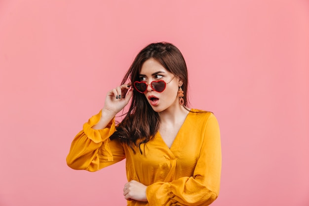 Foto gratuita dama elegante sorprendida se quita sus gafas en forma de corazón. foto de chica morena en blusa brillante en pared rosa aislada.