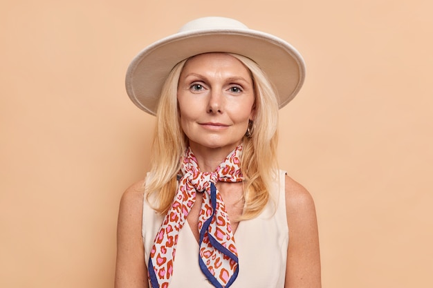 Dama femenina seria con maquillaje mínimo de cabello rubio vestida con un sombrero de camiseta blanca y un pañuelo atado alrededor del cuello que va a caminar posa en el interior contra la pared beige