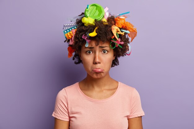 La dama insatisfecha preocupada se abrocha el labio inferior, recolecta basura plástica, usa una camiseta informal, es ecológica, molesta por un problema ambiental grave, tiene basura en el cabello rizado aislado en la pared púrpura