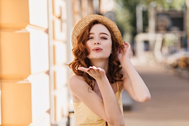 Dama de jengibre extática enviando beso de aire en la ciudad. Encantadora chica pelirroja con sombrero de verano relajante en buen día.