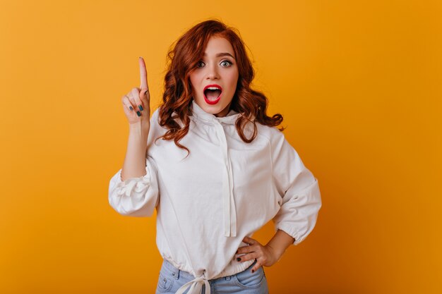 Dama de jengibre sorprendida con maquillaje elegante tiene una idea. Retrato de interior de glamorosa niña caucásica en suéter blanco.