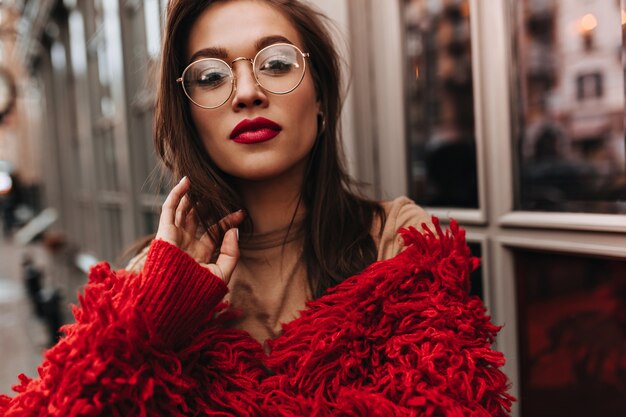 Dama con lápiz labial burdeos toca su cabello oscuro. Mujer de muy buen humor vestida con chaqueta de lana roja disfrutando de un cálido día de otoño afuera.