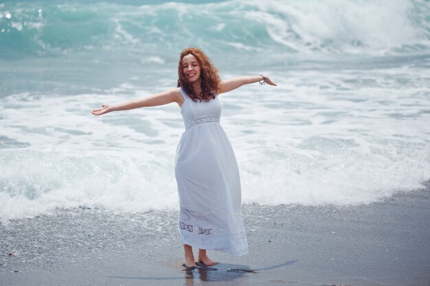 Dama de largo vestido blanco de pie con las manos abiertas en la playa durante el día