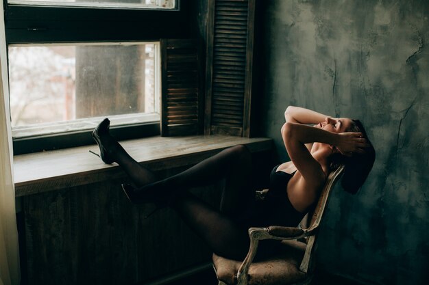 Dama en lencería negra y medias estira sus piernas en el alféizar de la ventana sentado en la silla