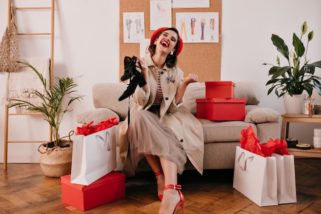 Dama de moda en boina roja y abrigo beige riendo y sosteniendo sandalias negras. Chica joven alegre con cabello oscuro disfruta de las compras.