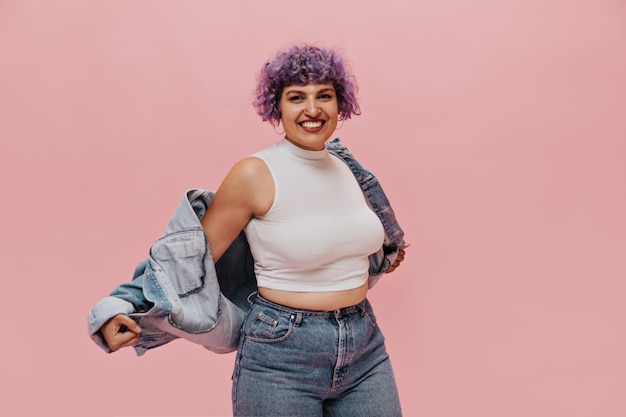 Dama de moda en jeans ajustados con estilo y top corto blanco en rosa. Mujer divertida con cabello violeta se quita la chaqueta.