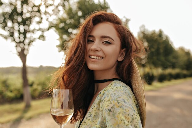 Dama de moda con peinado ondulado de jengibre y vendaje negro en el cuello con vestido verde moderno sonriendo y sosteniendo un vaso con bebida al aire libre