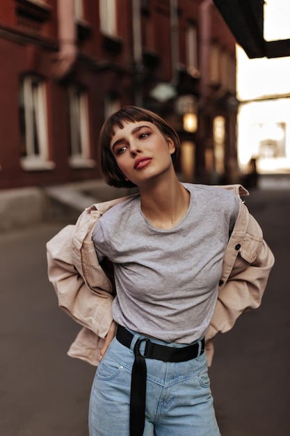 Dama de moda con el pelo corto posando en la ciudad Chica adolescente de moda en jeans de chaqueta de mezclilla beige con cinturón negro y tshort gris mirando hacia afuera