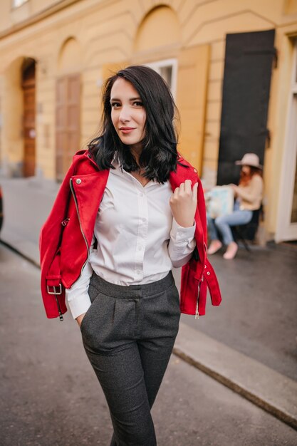 Dama morena delgada en pantalones grises posando con una sonrisa suave en la calle