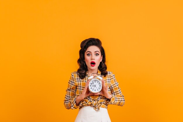 Dama morena sorprendida posando con reloj sobre fondo amarillo. Foto de estudio de chica pinup sorprendida viste camisa a cuadros.