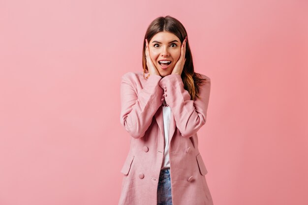 Dama morena sorprendida tocando la cara con una sonrisa. Disparo de estudio de maravillosa chica elegante aislada sobre fondo rosa.