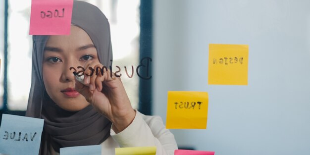 La dama musulmana asiática escribe información, estrategia, recordatorio en la placa de vidrio en la nueva oficina normal.