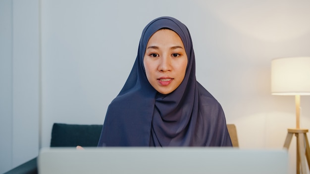 Foto gratuita dama musulmana usa hijab usando computadora portátil, hable con su colega sobre el plan en una reunión de videollamada mientras trabaja de forma remota desde casa por la noche en la sala de estar