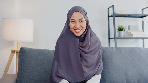 La dama musulmana usa hijab usando una computadora portátil, hable con sus colegas sobre el plan en una reunión de videollamada mientras trabaja de forma remota desde su casa en la sala de estar.