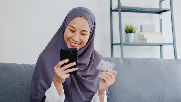 La dama musulmana usa un teléfono inteligente, compra con tarjeta de crédito y compra Internet de comercio electrónico en la sala de estar de la casa.