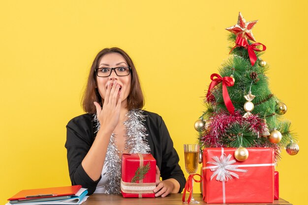 Dama de negocios emocional sorprendida en traje con gafas sosteniendo su regalo y sentada en una mesa con un árbol de Navidad en la oficina