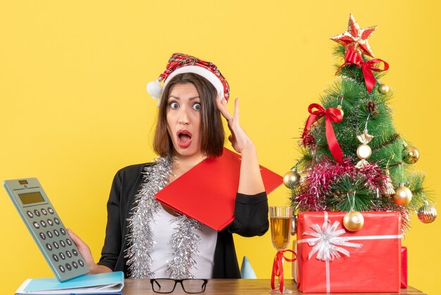 Dama de negocios sorprendida en traje con sombrero de santa claus y decoraciones de año nuevo sosteniendo calculadora y sentado en una mesa con un árbol de Navidad en la oficina