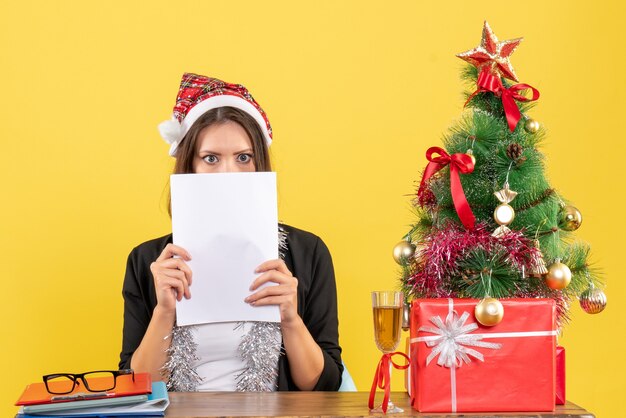 Dama de negocios sorprendida en traje con sombrero de santa claus y decoraciones de año nuevo trabajando solo y sentado en una mesa con un árbol de Navidad en la oficina