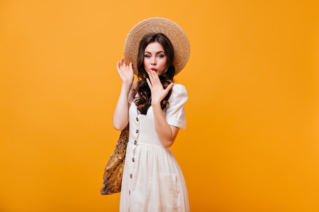 La dama de ojos verdes se cubre la boca con la mano. Mujer con sombrero de paja y vestido blanco tiene bolsa sobre fondo naranja.