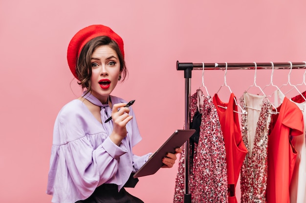 La dama de ojos verdes con sombrero rojo y labios rojos sostiene la tableta y mira a la cámara contra el fondo de perchas con ropa.