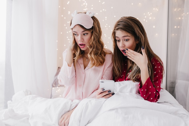 Dama de pelo largo sorprendida sentada bajo una manta blanca y mensaje de texto. Foto interior de chica rubia aburrida en máscara de dormir rosa.