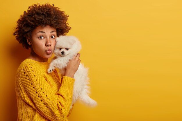 dama de piel oscura mantiene los labios redondeados, quiere besar a una adorable mascota, juega con un pequeño cachorro