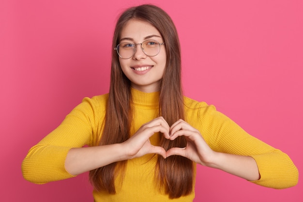 Foto gratuita la dama romántica viste una camisa casual amarilla que hace el símbolo del corazón con las manos, la mujer hace el signo de amor con los dedos