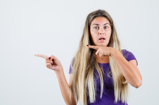 Dama rubia apuntando hacia un lado en camiseta violeta y mirando sorprendida. vista frontal.