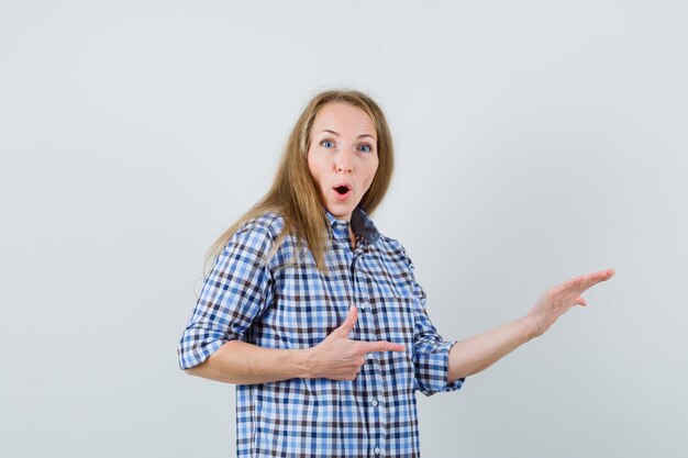 Dama rubia apuntando hacia un lado, levantando la palma en la camisa y mirando asombrada,